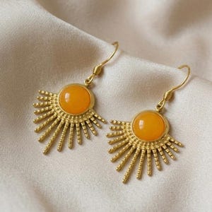 Könnte beinhalten: Ein Paar goldfarbene Ohrhänger mit einem zentralen, runden, orangefarbenen Cabochon-Stein. Die Metallfassung strahlt mit dünnen, texturierten Strahlen nach außen, die an ein Sonnenmotiv erinnern, und ist an einfachen goldfarbenen Ohrhaken befestigt.