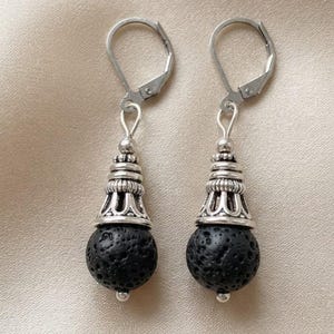 Può includere: Un paio di orecchini pendenti con perle sferiche di lava nera sospese a tappi in metallo argentato decorati e chiusure a leva. La consistenza porosa della pietra vulcanica scura contrasta con la finitura lucida della minuteria metallica.