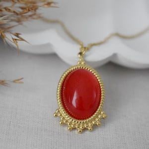 Könnte beinhalten: Eine goldfarbene Halskette mit einem ovalen Anhänger, der in der Mitte einen glatten, leuchtend roten Cabochon aufweist. Der Anhänger ist von einem gedrehten Seilrand umgeben und mit kleinen, abgerundeten Perlen am unteren Rand verziert.