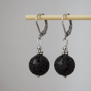 Op de afbeelding: Een paar oorhangers met zwarte lavasteen kralen. De oorbellen hebben zilverkleurige metalen accenten en een kleine zilveren kraal aan de onderkant van elke lavasteen bol. De oorbellen worden getoond tegen een neutrale achtergrond.
