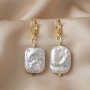 Könnte beinhalten: Ein Paar goldfarbene Ohrringe mit großen, unregelmäßigen, weißen, quadratisch geformten Perltropfen. Die Ohrringe haben einen Klappverschluss und eine kleine goldene Perle am unteren Ende jeder Perle. Der Hintergrund ist ein neutraler Satin-Stoff.