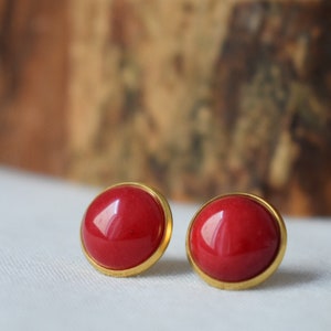 Peut inclure: Une paire de boucles d'oreilles rouges rondes avec une lunette dorée. Les boucles d'oreilles ont une finition brillante et sont posées sur un fond blanc doux. Ce sont des bijoux classiques.