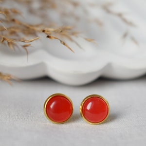 Può includere: Un paio di orecchini a bottone rotondi con un centro rosso brillante e un bordo dorato. Gli orecchini sono esposti su una superficie bianca con piante secche sullo sfondo. Un design di gioielli semplice ed elegante.