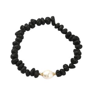 Könnte beinhalten: Schwarzes Lava-Gestein-Armband mit einer einzelnen weißen Perle und goldfarbenen Akzenten. Das Armband besteht aus kleinen, unregelmäßig geformten schwarzen Lavasteinen mit einer großen, glatten Perle in der Mitte.