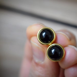 Può includere: Un paio di orecchini a bottone rotondi neri con un bordo color oro. Gli orecchini hanno una superficie lucida e riflettente. I gioielli sono tenuti in mano da una persona, con uno sfondo sfocato.