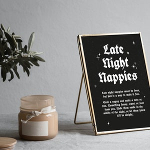 May include: A black framed sign with white text that reads "Late Night Nappies". The sign has a gold frame and is sitting on a table with a candle and a plant.