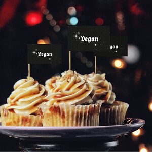 May include: Three vegan cupcakes with caramel frosting and pecan toppings, decorated with black flags that say "vegan" in white lettering.
