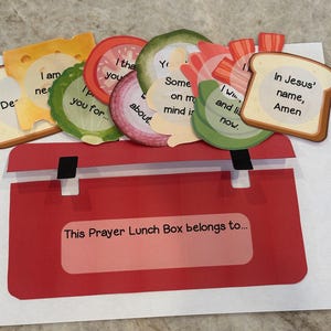 Könnte beinhalten: Eine rote Lunchbox mit einem weißen Etikett, auf dem "This Prayer Lunch Box belongs to..." steht. Die Lunchbox ist mit bunten Ausschnitten von Lebensmitteln verziert, auf denen jeweils ein anderer Spruch steht. Die Sprüche lauten: "I am needing you for...", "I thank you about...", "Some on my mind is...", "I will and listen now.", "In Jesus' name, Amen."
