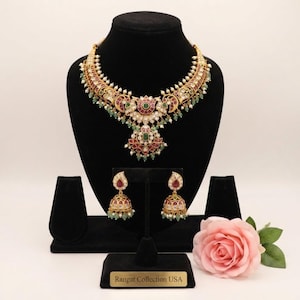 May include: A gold-toned necklace and matching jhumka earring set featuring intricate peacock motifs, inlaid with red and green stones, clear crystals, and small white pearls. The jewelry set includes a plaque labeled Rangat Collection USA.