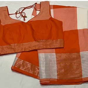 May include: A bright orange and white checkered saree with a matching orange blouse. The saree has a silver border with a striped pattern. The blouse has a V-neck and a silver trim.