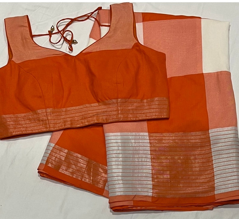 May include: A bright orange and white checkered saree with a matching orange blouse. The saree has a silver border with a striped pattern. The blouse has a V-neck and a silver trim.