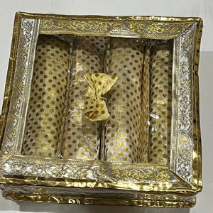 May include: A square storage box with a clear window lid, featuring a gold and silver metallic border with an ornate pattern. Inside, four cylindrical compartments are lined with cream fabric covered in a repeating gold polka dot pattern.