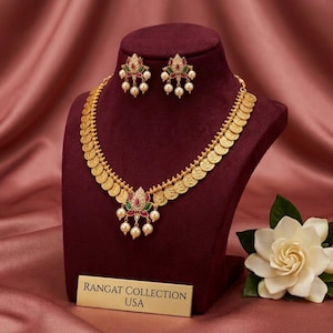 May include: Gold-toned necklace and earring set displayed on a maroon bust. The necklace features coin-shaped pendants and a central floral design with red, green, and white accents. The earrings mirror the floral design. A sign reads "Rangat Collection USA".