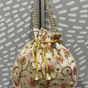 May include: A white drawstring bag with a pearl trim and a beaded strap. The bag is decorated with pink and gold embroidery of floral designs.