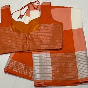 May include: Orange and white checkered saree with a matching orange blouse. The saree has a silver border and the blouse has a silver trim.