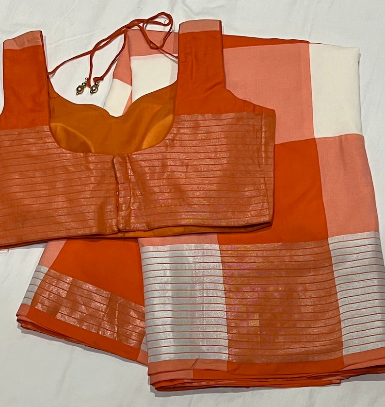 May include: Orange and white checkered saree with a matching orange blouse. The saree has a silver border and the blouse has a silver trim.