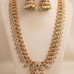 May include: A gold-toned necklace and matching jhumka earring set featuring a repeating paisley motif. The jewelry is embellished with white stones, red accents, and green teardrop-shaped stones, finished with dangling white and green beads.