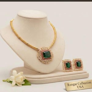 May include: A gold-toned necklace and matching square earring set featuring large emerald-green rectangular stones surrounded by small clear crystals and a thin border of red stones. A gold plaque reads Rangat Collection USA.