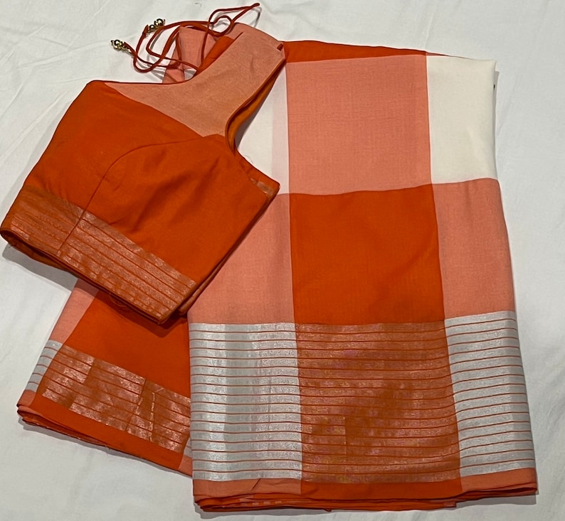 May include: A checkered orange and white saree with a matching orange blouse. The saree has a silver border with horizontal stripes.