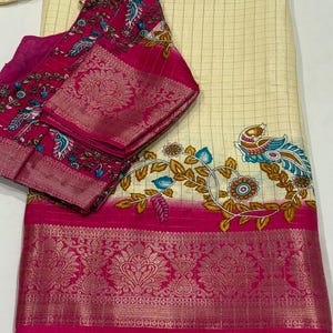 May include: A cream-colored saree with a checkered pattern and a pink border. The saree has a floral design with birds in blue, gold, and white. The saree comes with a matching pink blouse with a floral design.