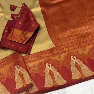 May include: A gold-colored saree with a red border featuring peacock and tree designs. A matching maroon blouse with gold accents is included. The saree has a textured pattern and is suitable for special occasions.