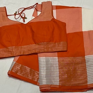 May include: A rust orange and white checkered saree with a matching rust orange blouse. The saree has a silver border with a striped pattern.