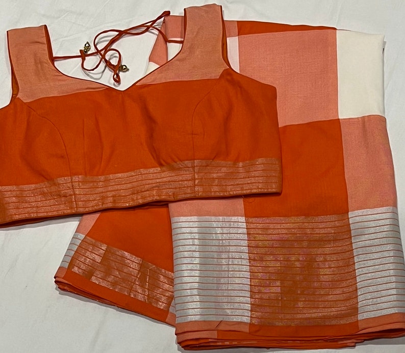 May include: A rust orange and white checkered saree with a matching rust orange blouse. The saree has a silver border with a striped pattern.