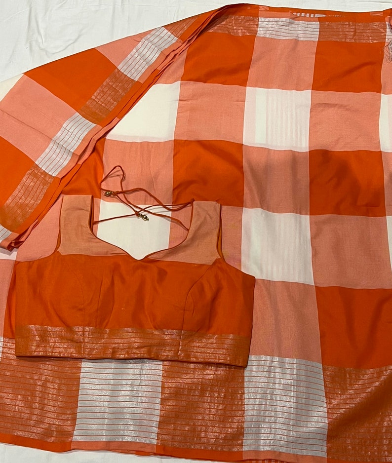 May include: A white and orange checkered saree with a matching orange blouse. The saree has a silver border and the blouse has a silver trim.