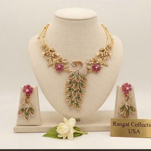 May include: A gold-toned necklace and earring set featuring a central peacock motif adorned with white, green, and pink stones. The necklace includes floral accents and a double-strand chain. A small gold plaque reads Rangat Collection USA.