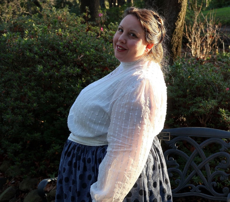 May include: A woman wearing a white, sheer blouse with a high collar and a blue polka dot skirt. She is standing in front of a bench with a leafy background.