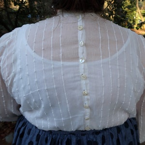 May include: A white sheer blouse with a button-up back. The blouse has a textured, dotted pattern and is made of a lightweight fabric.