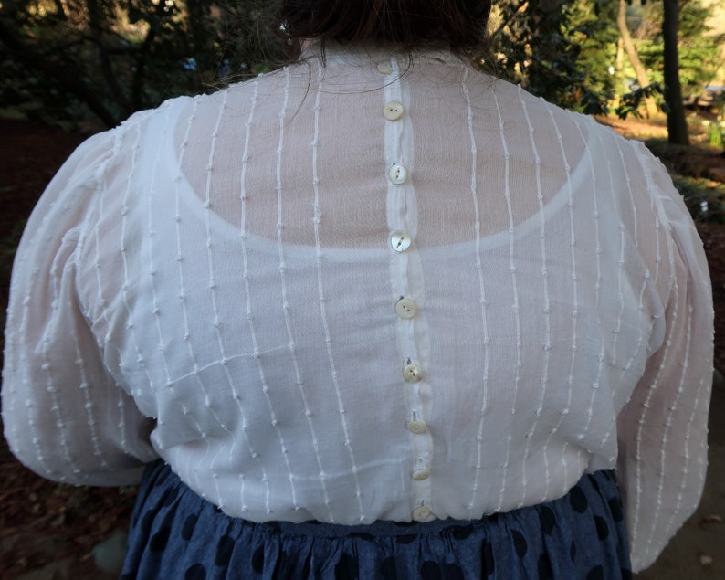 May include: A white sheer blouse with a button-up back. The blouse has a textured, dotted pattern and is made of a lightweight fabric.