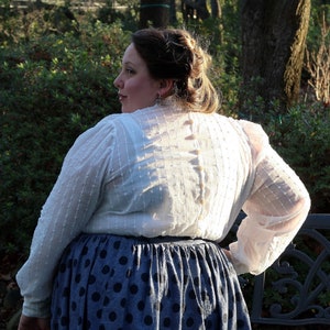 May include: A white, sheer blouse with a button-up back and a polka dot skirt. The blouse has a textured, almost quilted look.