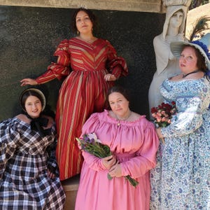 Peut inclure: Quatre femmes vêtues de robes d'époque posent en extérieur. L'une porte une robe rayée rouge et or, une autre une robe rose, une troisième une robe à fleurs bleues et la quatrième une robe à carreaux noirs et blancs. Une statue est en arrière-plan.