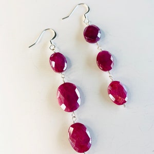 May include: A pair of dangle earrings with three oval-shaped, faceted, dark red gemstones on each earring. The earrings are made with silver-toned metal.