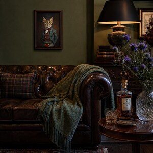 May include: A framed portrait of a cat dressed in a plaid suit and bowtie hangs above a dark brown tufted leather sofa. The sofa is draped with a green fringed throw blanket and a plaid pillow. A side table holds a glass of amber liquid.