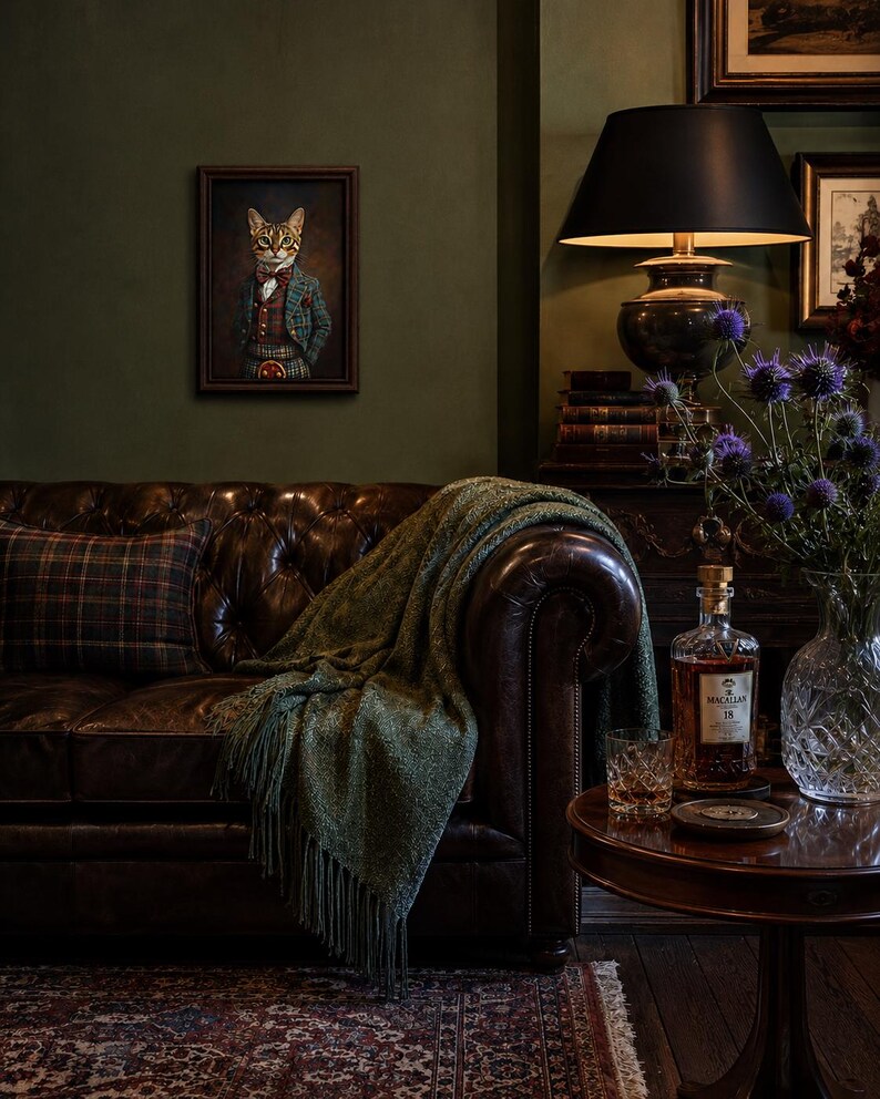 May include: A framed portrait of a cat dressed in a plaid suit and bowtie hangs above a dark brown tufted leather sofa. The sofa is draped with a green fringed throw blanket and a plaid pillow. A side table holds a glass of amber liquid.