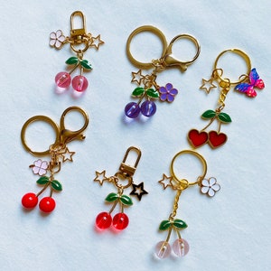 May include: Six gold keychains with different charms. Each keychain features a cherry design with a different colored glass cherry. The charms include a pink flower, a purple flower, a red heart, a butterfly, a star, and a clover.