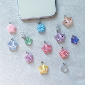May include: A selection of colourful star-shaped phone charms in shades like pink, blue, green, and yellow. Some charms feature smiling faces. The charms are attached to a phone and are on a white surface.
