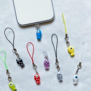 Puede incluir: Una colección de ocho abalorios de teléfono con forma de pez de colores con ojos negros y lunares blancos. Cada abalorio está unido a una cuerda y un anillo de metal.