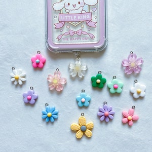 Puede incluir: Una funda transparente para teléfono con borde rosa y un diseño de perro de dibujos animados, junto con una colección de colgantes de flores de colores. Los colgantes son de varios colores, incluyendo rosa, azul y amarillo, y tienen pequeños bucles metálicos.