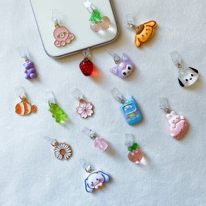 Puede incluir: Una colección de 14 charms de teléfono coloridos con varios diseños, incluyendo una fresa, un melocotón, una flor, un pez, un osito de goma, un perro, un gato, un teléfono, un conejito y un pulpo.