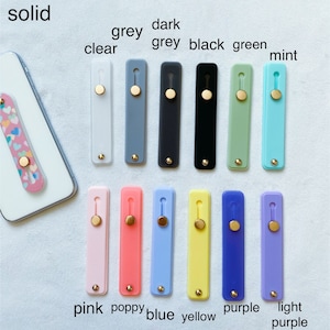 May include: A variety of phone grips in different colors, including clear, grey, dark grey, black, green, mint, pink, poppy, blue, yellow, purple, and light purple. The grips are all made of silicone and have a gold button.