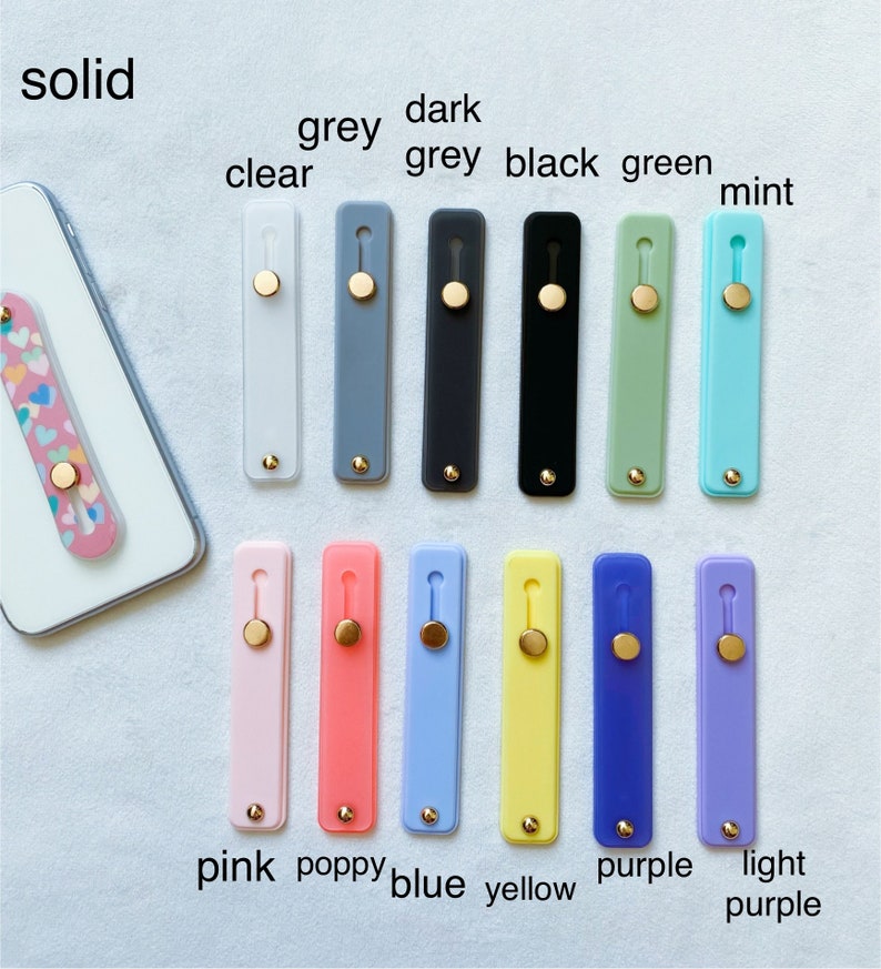 May include: A variety of phone grips in different colors, including clear, grey, dark grey, black, green, mint, pink, poppy, blue, yellow, purple, and light purple. The grips are all made of silicone and have a gold button.