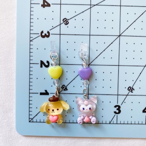 May include: Two charm keychains with pastel coloured characters. The keychain on the left features a yellow dog with a brown hat and a pink heart. The keychain on the right features a purple character with a pink heart and a white face.