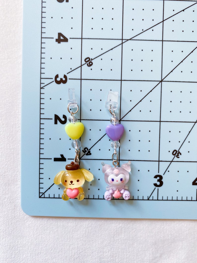 May include: Two charm keychains with pastel coloured characters. The keychain on the left features a yellow dog with a brown hat and a pink heart. The keychain on the right features a purple character with a pink heart and a white face.