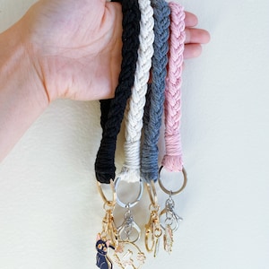 Op de afbeelding: Gevlochten sleutelhangers in zwart, crème, grijs en roze. Elke sleutelhanger heeft een goud- of zilverkleurige ring en sluiting, met decoratieve bedels, waaronder een zwarte kat en bloemmotieven.
