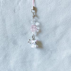 May include: A pink phone charm featuring a white cat figurine with pink accents, a silver star charm, and clear and pink beads. The charm is attached to a pink cord with silver hardware.
