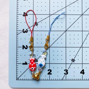May include: Two phone charms with red and blue fish charms, gold beads, and a gold heart charm. The charms are attached to red and blue strings.