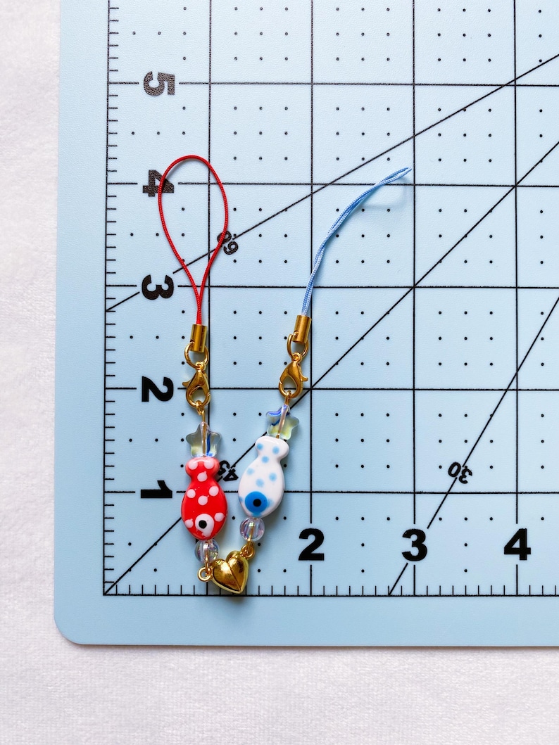 May include: Two phone charms with red and blue fish charms, gold beads, and a gold heart charm. The charms are attached to red and blue strings.